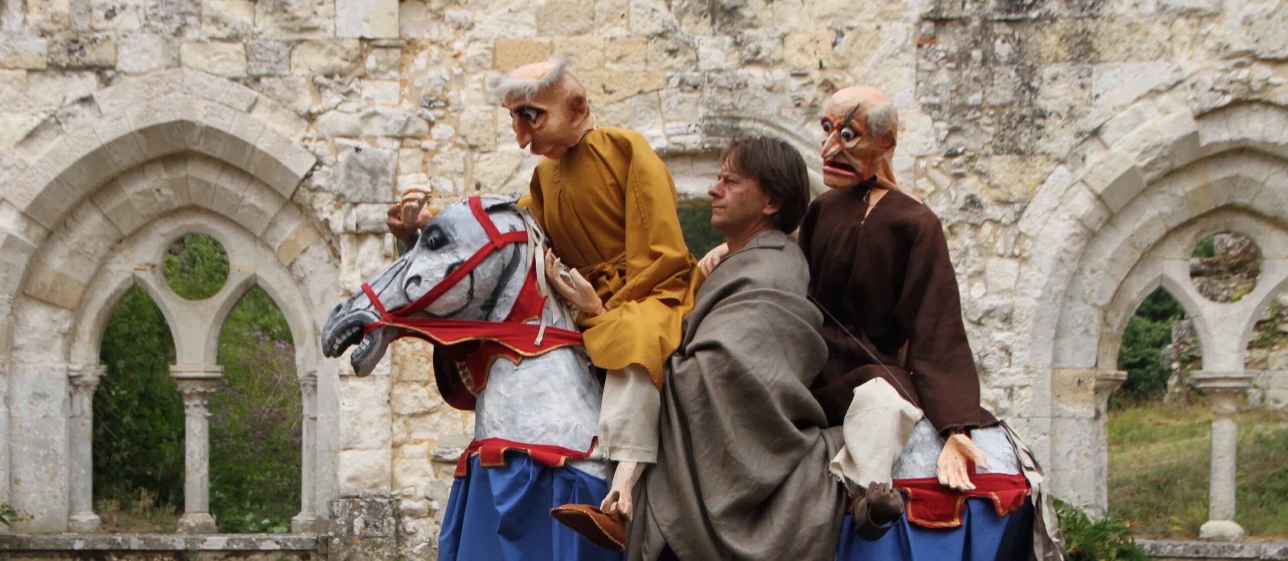 Deux individus portant des masques surdimensionnés et des vêtements médiévaux se produisant sur un faux cheval dans un contexte historique.