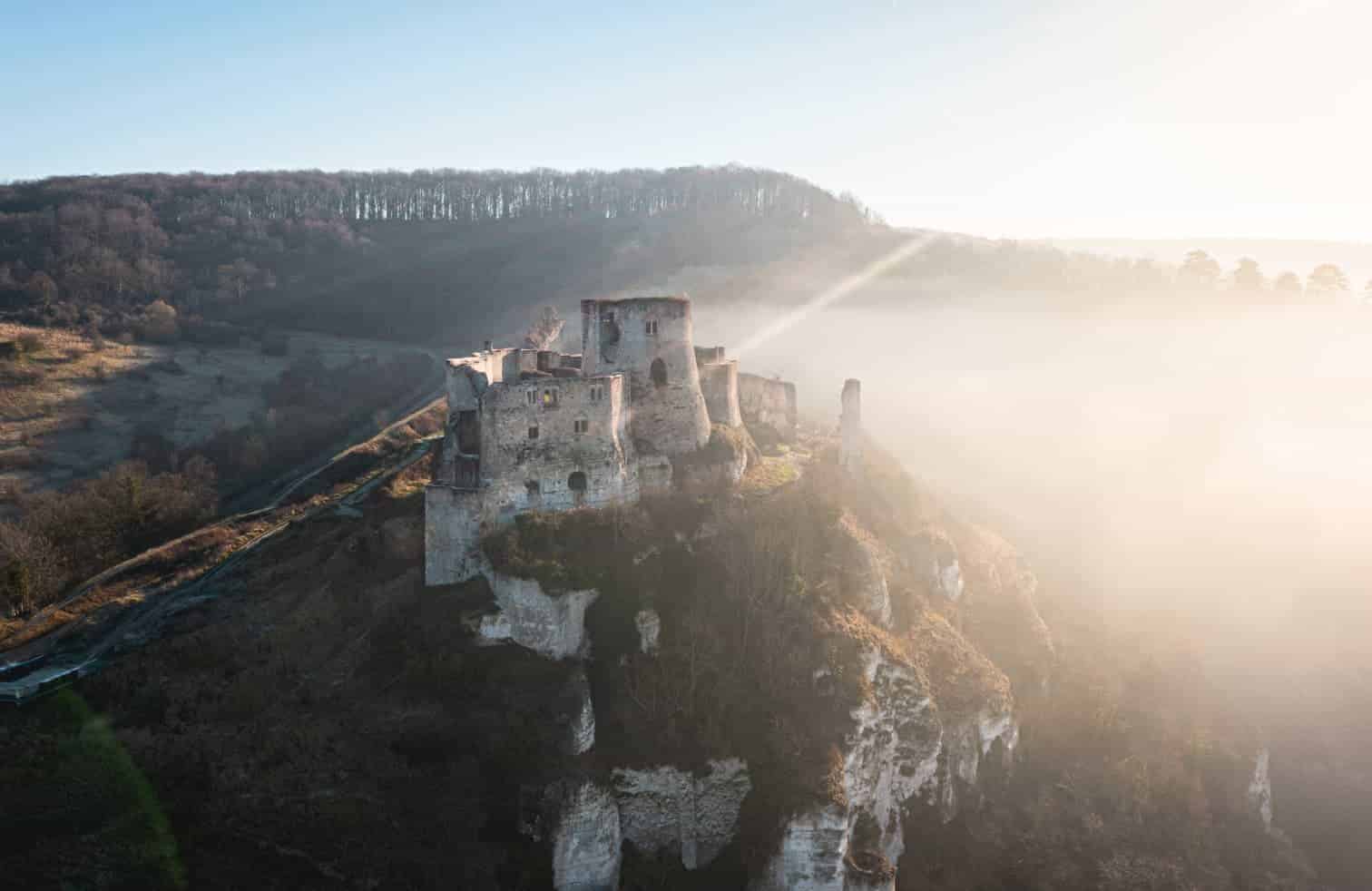 Ancien château perché sur une falaise surplombant une vallée couverte de brume au lever du soleil.