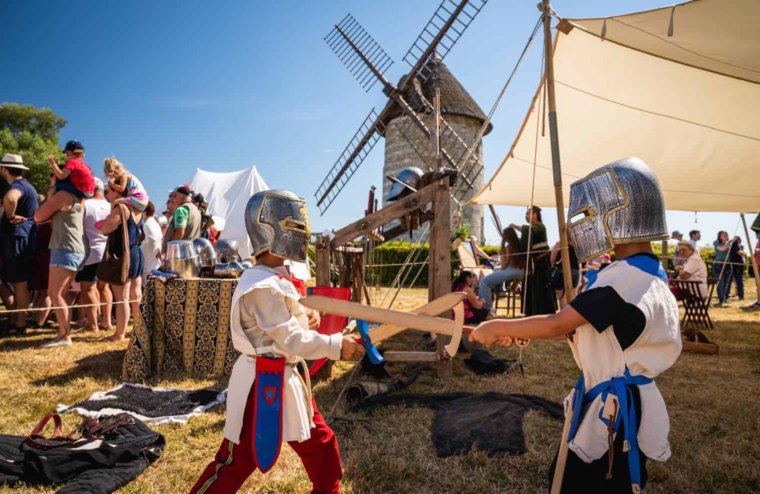 Deux enfants en armure médiévale jouent au combat avec des épées en bois lors d'une reconstitution historique en plein air, avec un moulin à vent en arrière-plan et des spectateurs observant.
