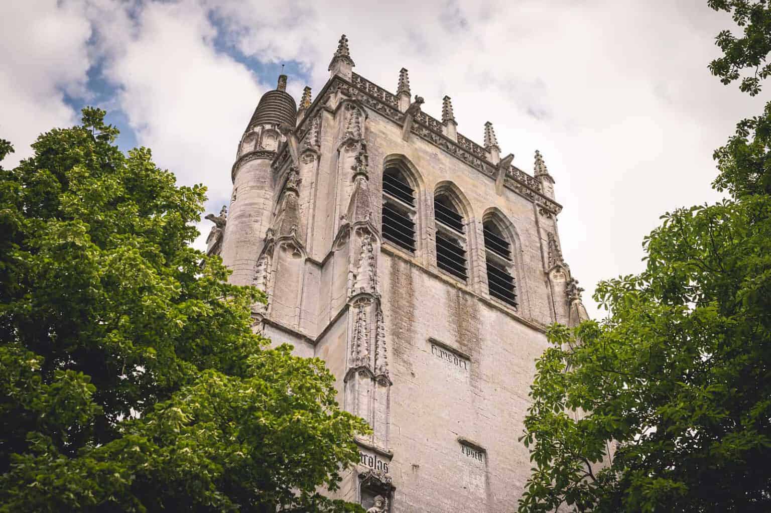 Tour en pierre historique à l'architecture gothique surplombant des arbres verdoyants contre un ciel nuageux.