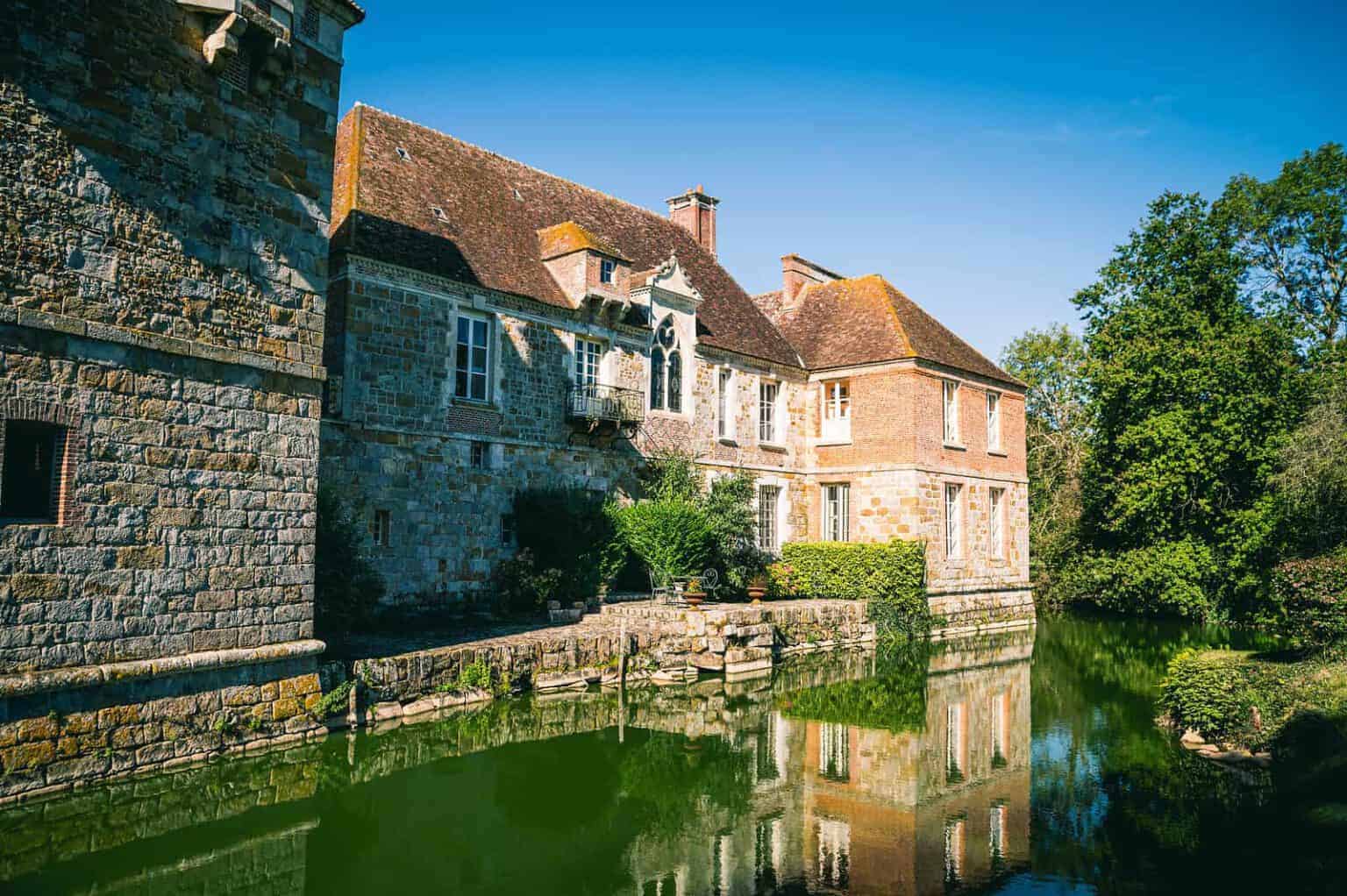 Maison traditionnelle en pierre avec un toit de tuiles rouges au bord d'un étang calme par temps clair.