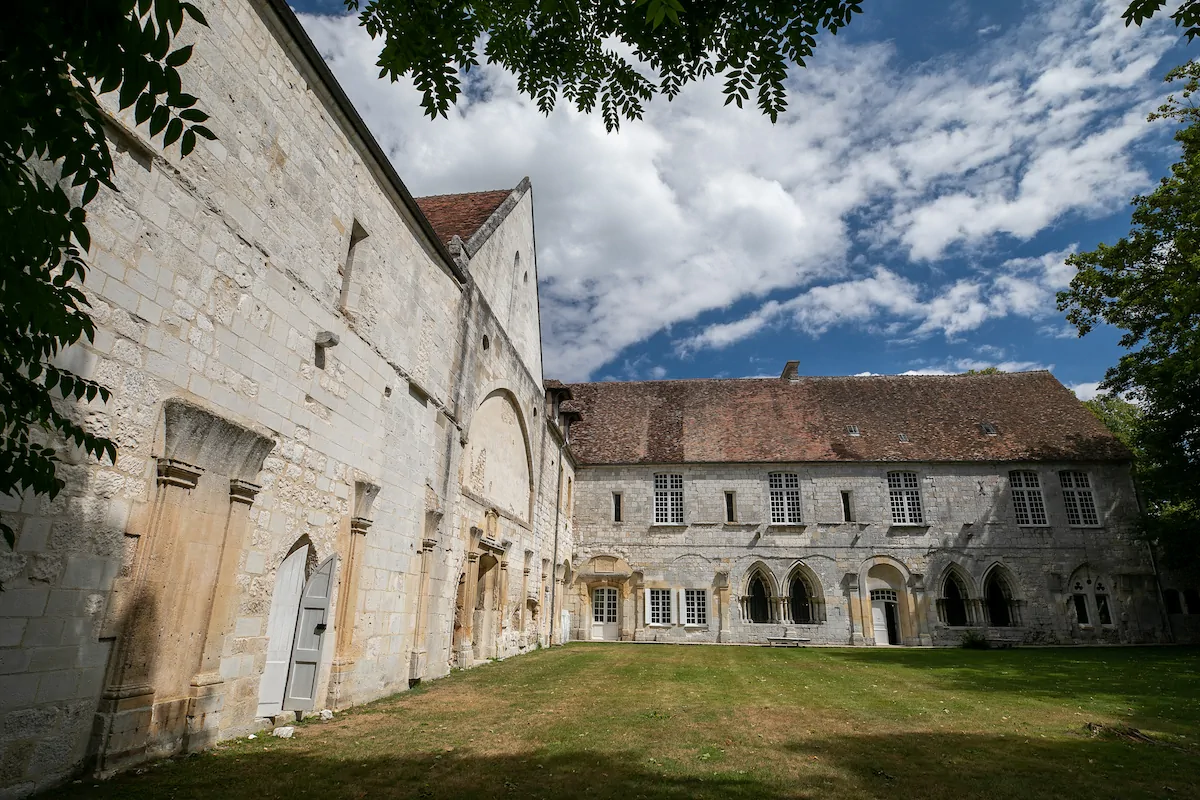 Un bâtiment historique en pierre avec des fenêtres cintrées forme une cour avec une pelouse verdoyante à l'Abbaye de Bonport. Le bâtiment présente un mélange de pierres beiges et grises et un toit de tuiles rouges. Le ciel bleu vif avec des nuages épars encadre la scène, avec des branches d'arbres suspendues au sommet.