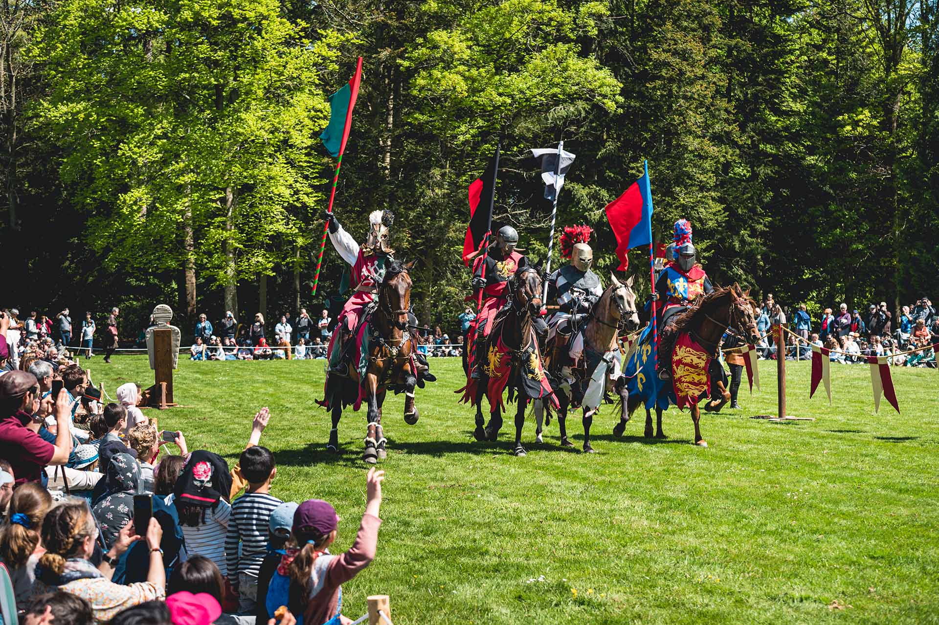 Quatre chevaliers médiévaux en armure colorée montent à cheval, portant des drapeaux, vers une foule assise sur l'herbe lors d'une foire de la Renaissance ou d'une joute. Le décor d’arbres verts luxuriants rehausse l’authenticité de la scène du Moyen Âge. Les spectateurs, y compris les enfants et les adultes, regardent avec impatience et saluent les chevaliers.